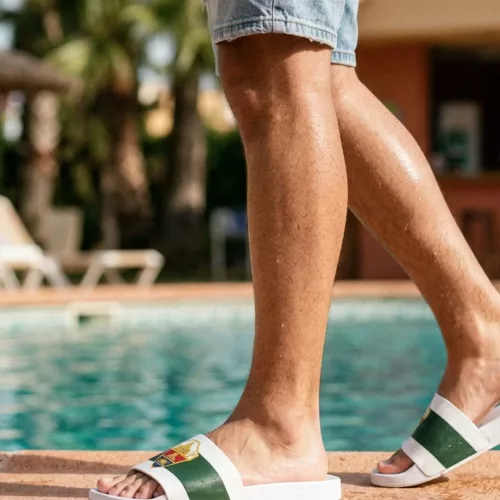 Vista lateral de las piernas de un hombre caminando por el borde de una piscina, usando chanclas blancas oficiales del Elche CF con franja verde y escudo, con palmeras de fondo en un día soleado.