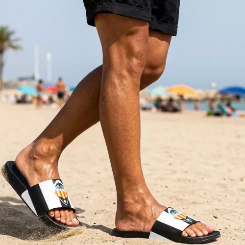 Un modelo masculino con pantalones cortos negros camina por la arena de una playa soleada, usando chanclas oficiales del Valencia CF de color negro con franjas blancas y el escudo del equipo en la tira