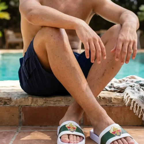 Un hombre sentado en el borde de una piscina de piedra, con las piernas cruzadas y el torso desnudo, usa chanclas blancas oficiales del Elche CF con franja verde y escudo del club, en un entorno de verano.