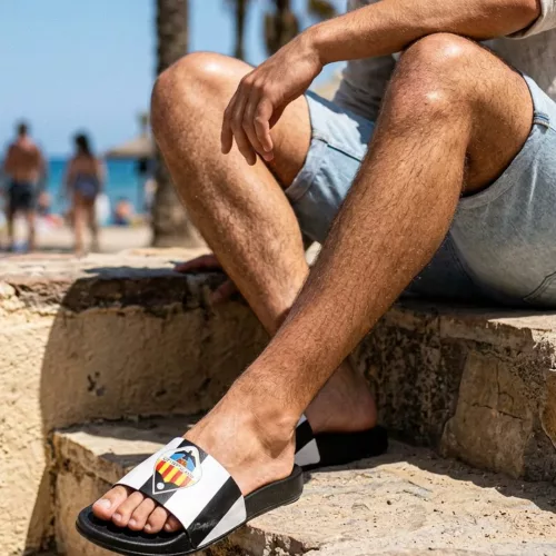Primer plano de las piernas de un hombre sentado en escaleras de piedra en la playa, usando chanclas negras oficiales del Valencia CF con franjas blancas y escudo, con el mar desenfocado al fondo.