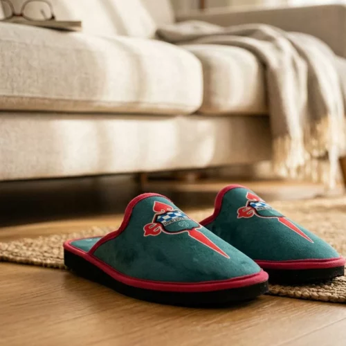 Detalle de zapatillas de estar por casa del Albacete Balompié con escudo bordado, usadas por un hombre con pantalones vaqueros doblados en un ambiente de luz natural.