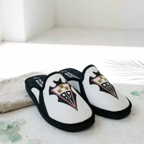 Primer plano de una persona caminando con zapatillas de casa oficiales del RC Celta de Vigo, color verde con ribete rojo, en un salón acogedor.