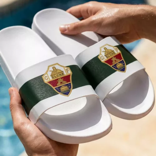 Primer plano de unas manos sosteniendo un par de chanclas oficiales del Elche CF, de color blanco con una franja verde central y el escudo del club en detalle, con una piscina desenfocada de fondo.